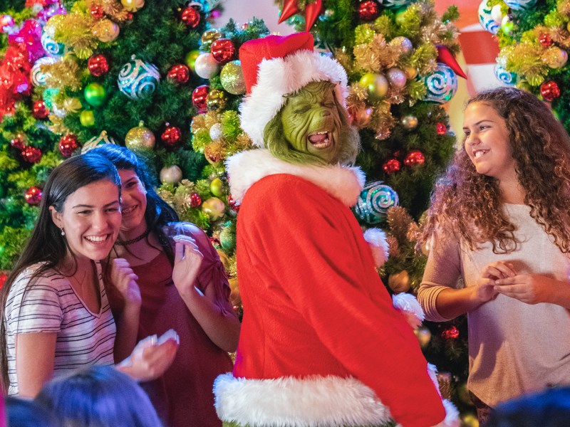 The Grinch, dressed in a red Santa suit, stands in the center smiling with his mouth open, interacting with three young women who are all smiling at him. They are surrounded by heavily decorated and brightly lit Christmas trees.
