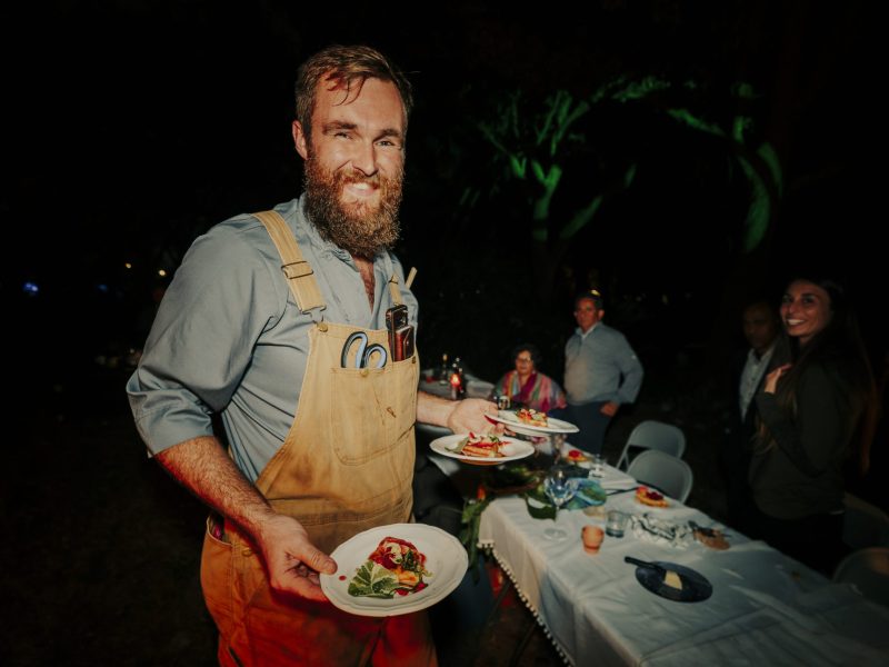 A bearded man wearing tan overalls over a blue shirt smiles broadly while balancing three plates of food on his arm and hand. He has a pair of scissors tucked into his chest pocket and stands in front of a long dining table set for an outdoor evening event, with other guests visible in the dark background.