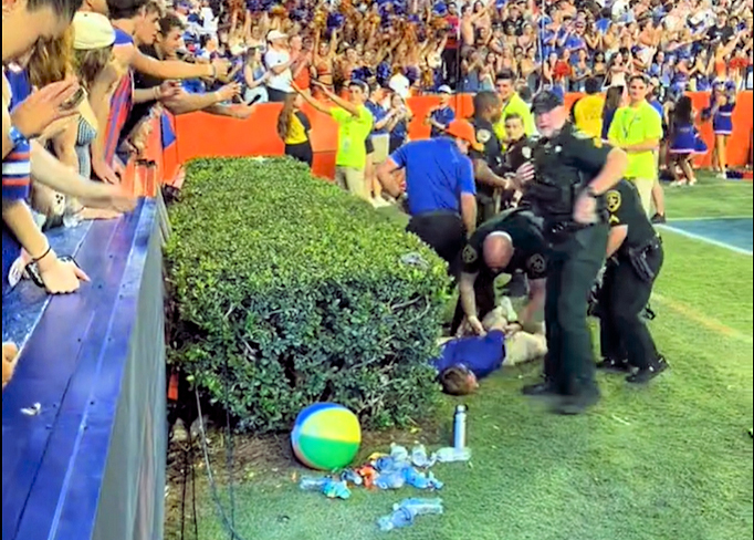Several police officers and security personnel restraining a person on the ground near the stands and a hedge on a crowded football field; spectators watch from above.