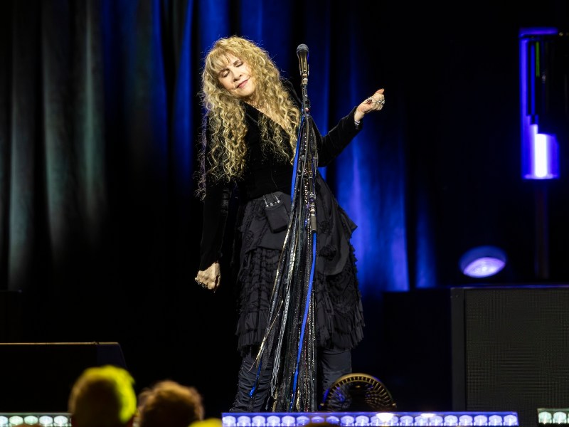 A performer with long curly blonde hair stands on stage wearing a flowing black outfit, eyes closed and hands raised slightly as she sings into a microphone draped with scarves. Blue and purple stage lighting illuminates the dark background.