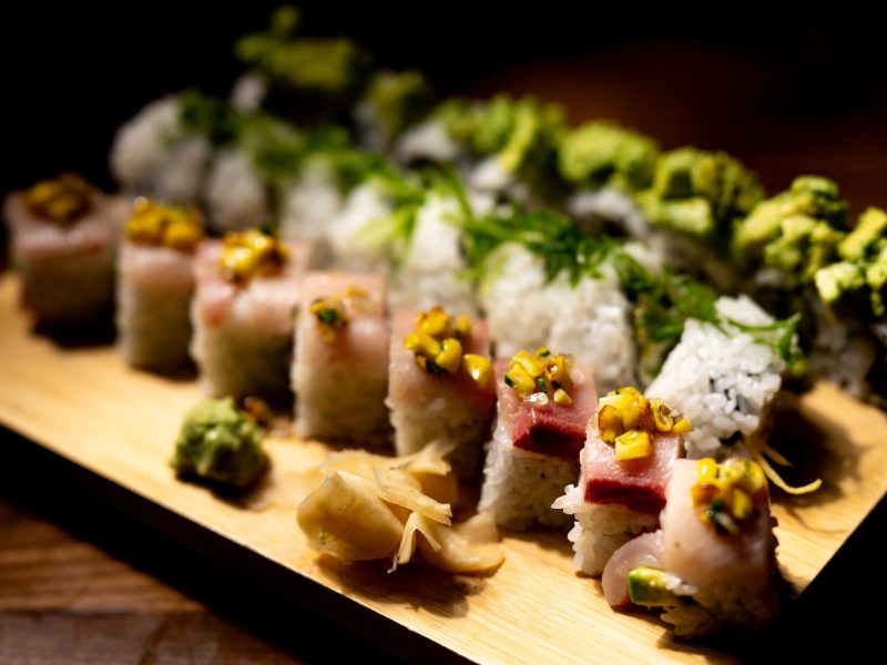 A rectangular wooden board displays several rows of sushi rolls, dramatically lit against a dark background. The front row features rolls topped with slices of pink raw fish and a yellow corn relish or garnish, while the back rows include rolls topped with avocado and herbs. A pile of pickled ginger and a dollop of wasabi sit on the board in the foreground.