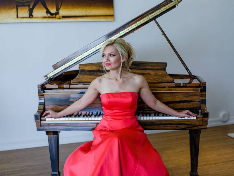 A woman with blonde hair, wearing a vibrant red strapless dress, sits gracefully at a grand piano. Her hands are resting on the keys, and she is looking to her left with a pensive expression. The piano is made of wood with a striking striped grain. In the background, there is a large, horizontal painting on a white wall that depicts a person's legs in high heels. A faint glimpse of a rug is also visible on the floor to the far right.