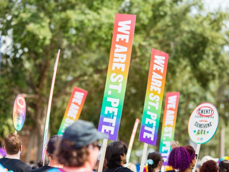 A crowd of people, viewed from behind, holds up several tall, rectangular signs featuring a rainbow gradient and the text "WE ARE ST. PETE" in white block letters. To the right, a circular sign reads "Twenty Years of Pride in the Sunshine City," all set against a backdrop of green trees.