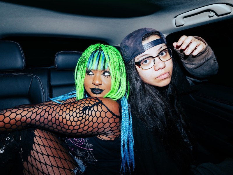 Two members of Meet Me @ The Altar posing in the back seat of a car at night. One has vibrant neon green and blue braids, dark makeup, a nose ring, and is wearing fishnets. The other has dark hair, glasses, a lip ring, and a backwards baseball cap.