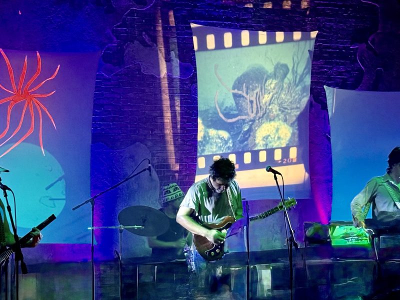 A band performs live on stage under blue and purple lighting, with a backdrop displaying film-strip projections and abstract red spider-like artwork.