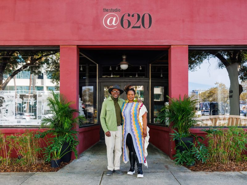 A man in a green shirt and hat, and a woman in a long, brightly colored striped dress, stand smiling in front of the red facade of "The Studio @ 620" venue.