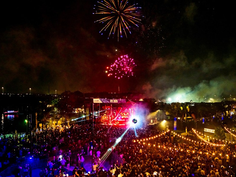 A vibrant night scene captures a massive crowd gathered for an outdoor concert and celebration. Above the stage, which is bathed in red light and marked with a banner reading "NYE 2025," colorful fireworks explode in the dark sky. String lights drape over the audience on the right, while the water on the left reflects the blue and green ambient lights of the venue.