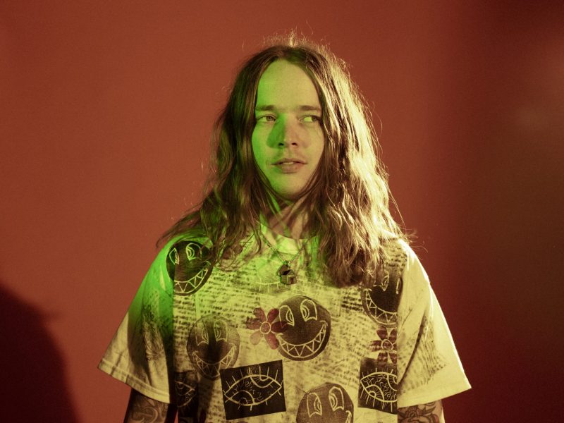 A person with shoulder-length wavy hair and a neutral expression looks off to the right. The subject is wearing a printed t-shirt featuring smiley faces and eyes, with tattoos visible on the arms, under harsh studio lighting.