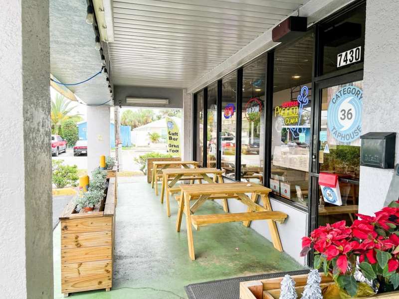 The exterior of "Category 36 Taphouse" features a row of wooden picnic tables arranged on a green painted patio under a covered walkway. Large storefront windows display neon beer signs, such as Blue Point and Pacifico, next to a glass door labeled with the address "7430" and the business logo. Wooden planters with greenery border the left side, while red poinsettias add festive color to the foreground.