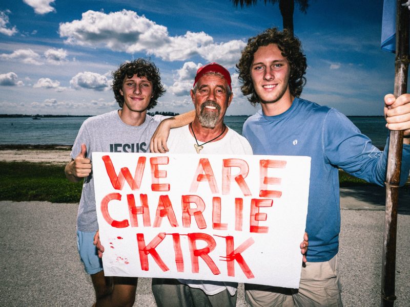 A man in a red hat stands between two young men with curly hair. They are holding a large white sign with the phrase "WE ARE CHARLIE KIRK" written on it in bold, red paint. They are standing near a body of water under a partly cloudy sky.