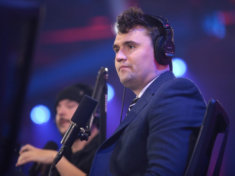 A man with dark hair and a suit A man in a dark suit jacket and tie is seated in a dark, studio-like setting, wearing a pair of black over-ear headphones. He is looking off to the side with a serious expression. A microphone on an arm stand is positioned in front of him, and another person in a beanie is blurred in the background.