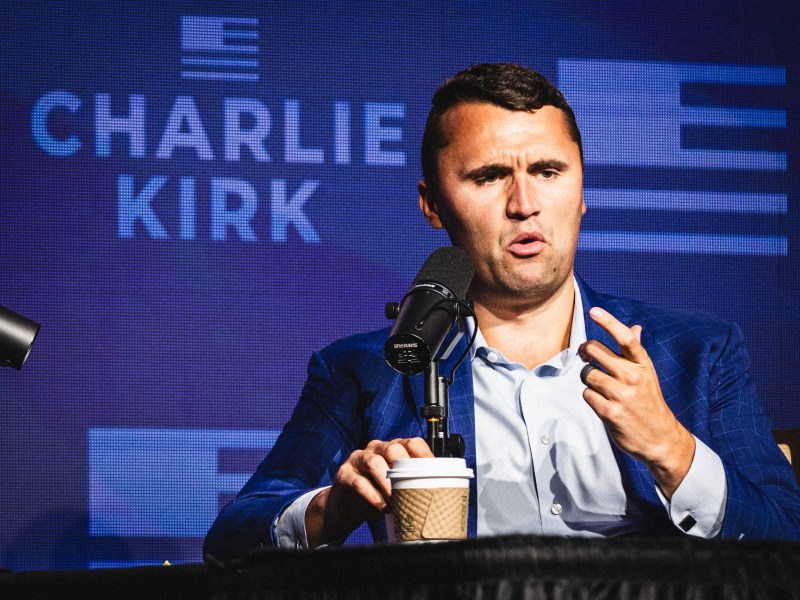 Close-up of Charlie Kirk seated at a panel, speaking into a microphone and gesturing with his right hand, wearing a blue patterned suit. A coffee cup rests on the table in front of him, and his name is visible on the blue backdrop.