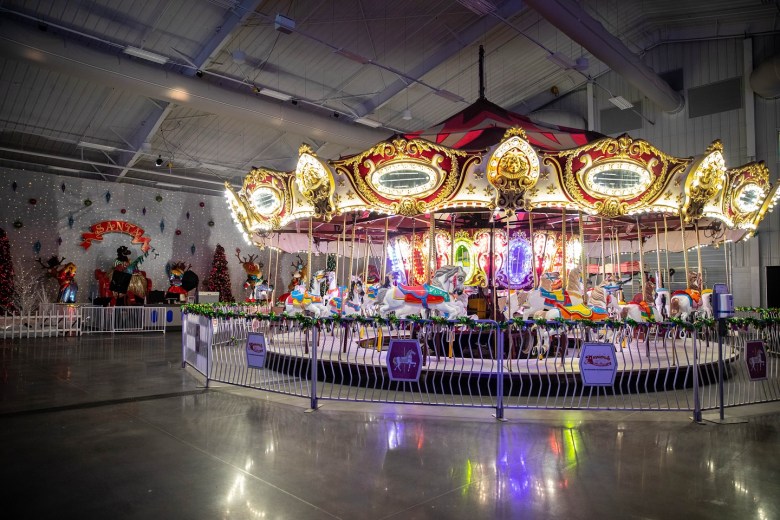 A brightly illuminated, ornate carousel featuring gold detailing and colorful horses sits in the middle of a large warehouse-style room with polished floors. In the background to the left, a festive holiday section is visible, complete with a "SANTA" sign, decorated trees, and animatronic figures, all enclosed by a low white metal fence.