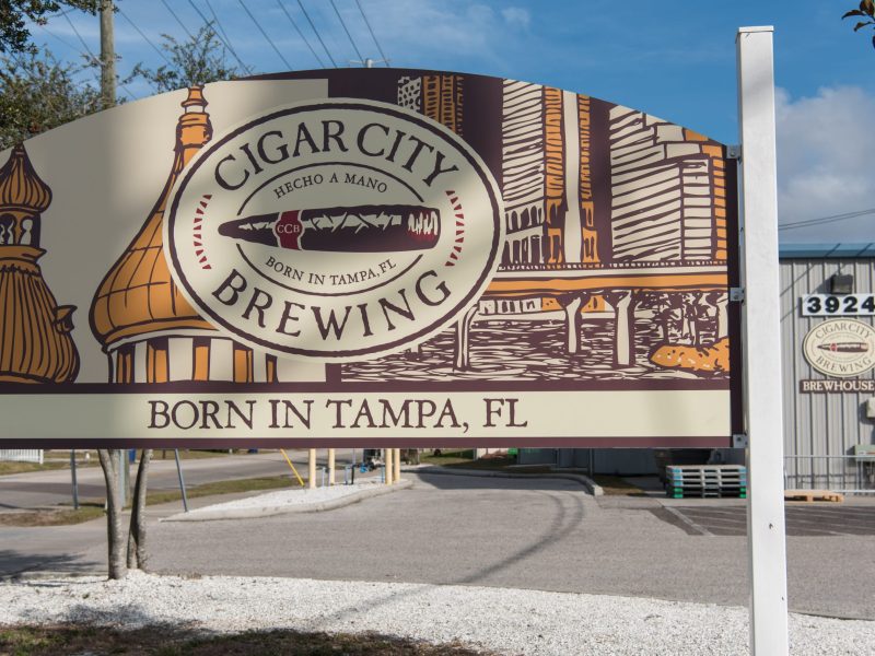 Outdoor sign for Cigar City Brewing featuring a logo with a cigar graphic and the text "BORN IN TAMPA, FL" over a background illustration of buildings. The brewery entrance is visible in the background.