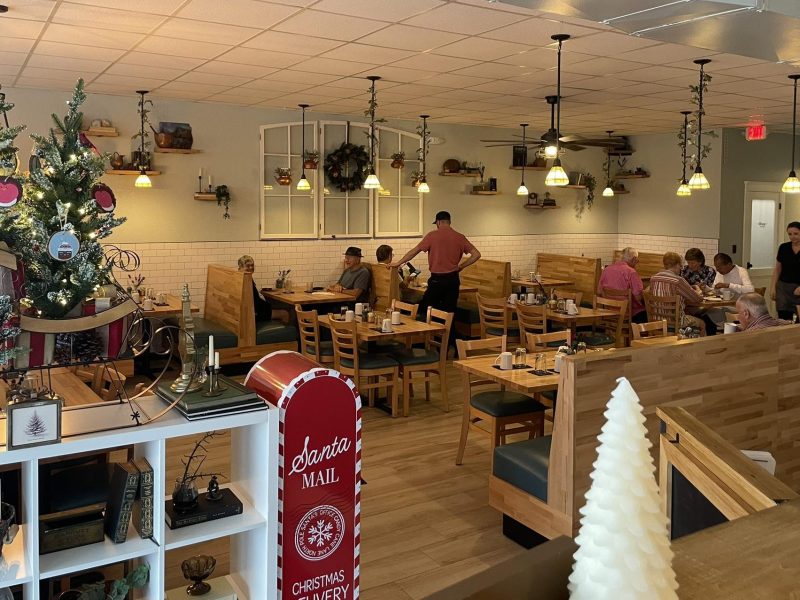 The dining room of a restaurant is decorated for the holidays, featuring wooden tables and booths where patrons are seated. A server in a red shirt stands near the center attending to customers, while the room is lit by pendant lights wrapped in greenery. Festive touches include a Christmas tree with ornaments, a red "Santa MAIL" box in the foreground, and wreaths hanging on decorative wall window frames.
