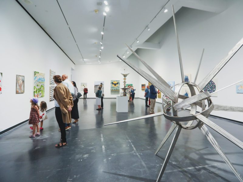 A large, metallic starburst sculpture stands in the foreground of a bright art gallery while visitors view paintings in the background.