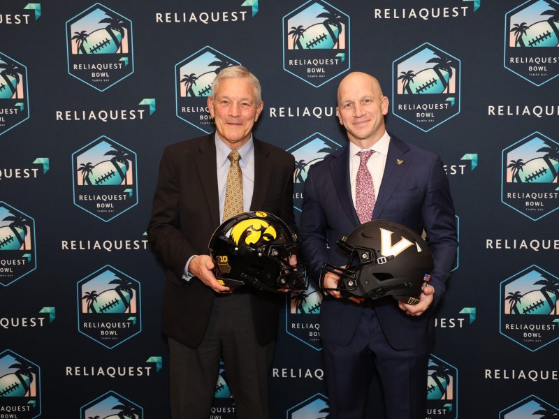 Two smiling men in formal business suits stand side-by-side against a backdrop featuring repeated "ReliaQuest Bowl Tampa Bay" logos. The man on the left holds a black football helmet with a yellow hawk logo, while the man on the right holds a black football helmet with a white "V" logo.