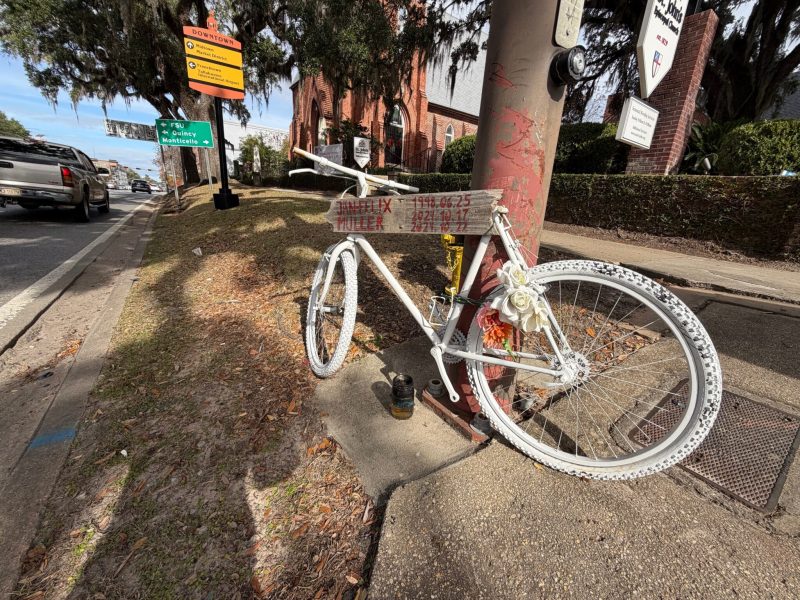 A roadside memorial featuring a bicycle painted entirely white, known as a "ghost bike," chained to a utility pole on a sunny sidewalk. A wooden sign attached to the frame displays the handwritten name "Jan Felix Muller" along with dates ranging from 1998 to 2024. Artificial white and peach-colored flowers decorate the rear wheel, and the background shows a street corner with signs pointing toward "Downtown," "FSU," and the "Tallahassee International Airport," alongside a brick church.