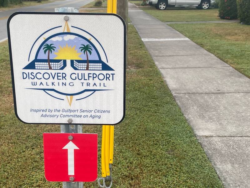 A square sign for the "Discover Gulfport Walking Trail" featuring a stylized logo with palm trees and a compass rose. Below it, a small red sign with a white arrow points straight up, indicating the trail direction.
