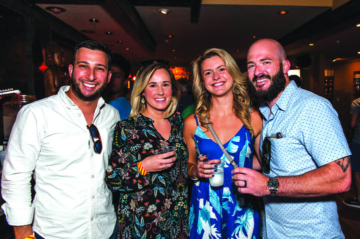 A waist-up, indoor shot of four people standing side-by-side and smiling at the camera, likely at a bar or social event. From left to right: a man in a white button-down shirt, a woman in a dark floral dress, a woman in a blue and white patterned dress, and a bald man with a beard in a light blue patterned shirt.