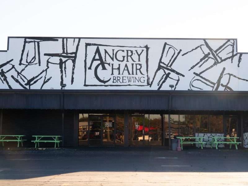 The exterior of the Angry Chair Brewing building features a distinctive mural above the awning, depicting abstract black sketches of chairs against a white background. Below the dark awning, the storefront has large tinted windows and glass entrance doors. Several bright green picnic tables are arranged on the pavement in front of the building.