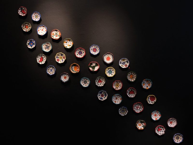 A display of small, colorful ceramic bowls mounted on a black wall, arranged in a diagonal, wave-like formation from the top left to the bottom right. Each bowl features unique hand-painted designs, including floral patterns, geometric shapes, and fruit motifs.