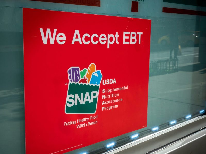 A red sign posted on a store window reads "We Accept EBT" and features the SNAP (Supplemental Nutrition Assistance Program) logo with the text "Putting Healthy Food Within Reach."
