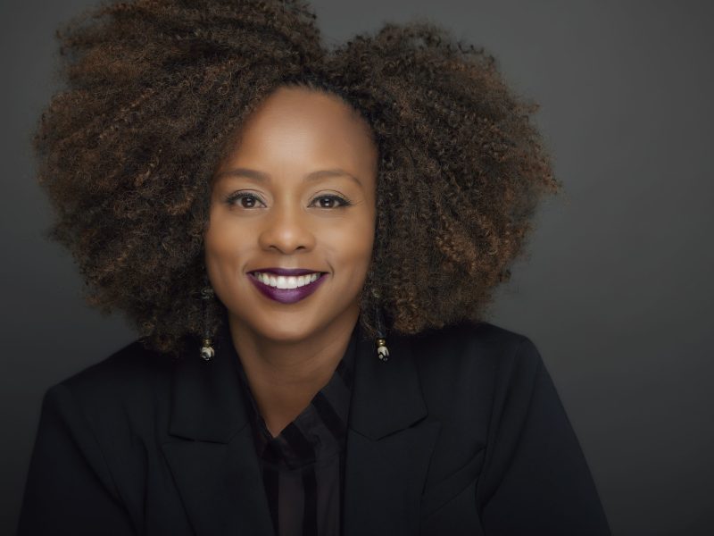 Professional headshot of a smiling Black woman with a large, textured afro, wearing a black blazer and dark purple lipstick, against a gray background.