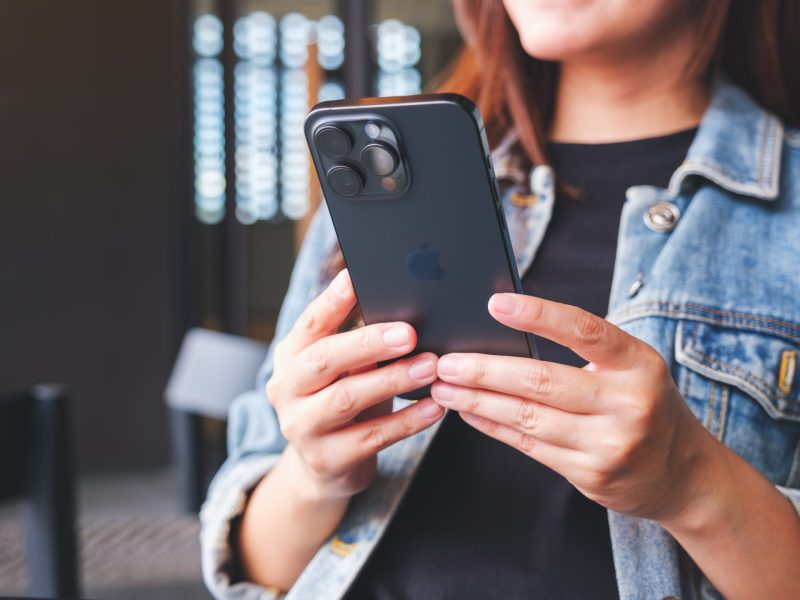 Close-up of a person wearing a denim jacket, holding a dark-colored iPhone with the triple camera system visible on the back.