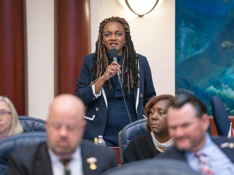 A professional photo of an African American woman at a legislative session or meeting, holding a microphone and addressing the room. She has an engaged expression. In the background is a large abstract blue painting, and seated participants are blurred in the foreground.