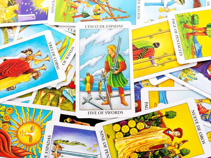 The Five of Swords tarot card from the Rider-Waite deck is centered among a pile of scattered cards. The card depicts a man holding three swords and looking over his shoulder while two defeated figures walk away, leaving two swords on the ground.