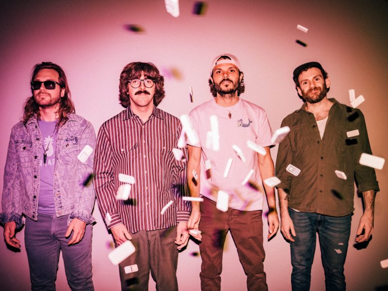 Portrait of the four members of the band Goose standing against a pink backdrop as white confetti or papers fall around them; Photo Credit: Juliana Bernstein.