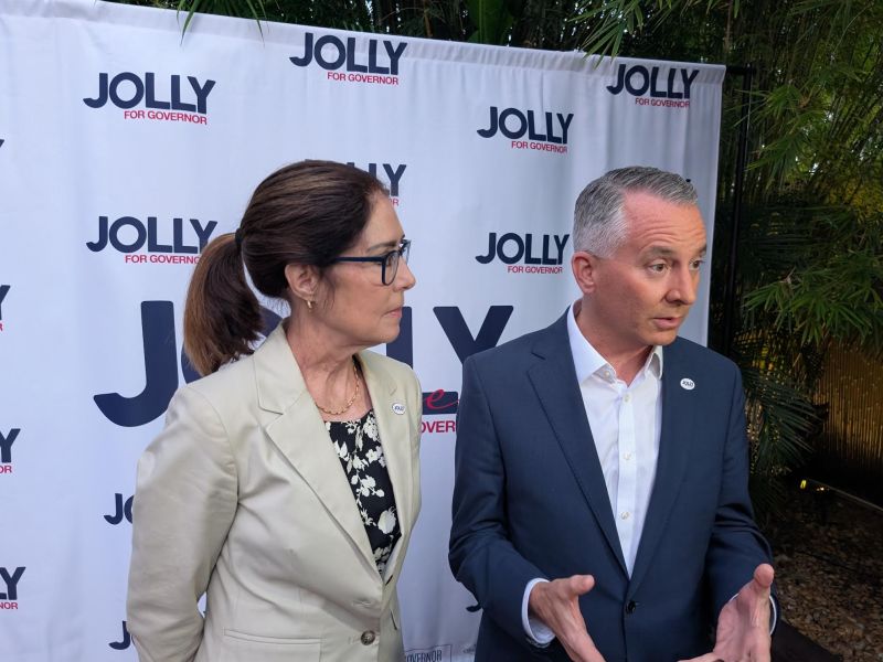 A close-up shot of two people engaged in conversation or an interview at a political rally. One individual is speaking emphatically while the other is listening intently, framed by large campaign signs.