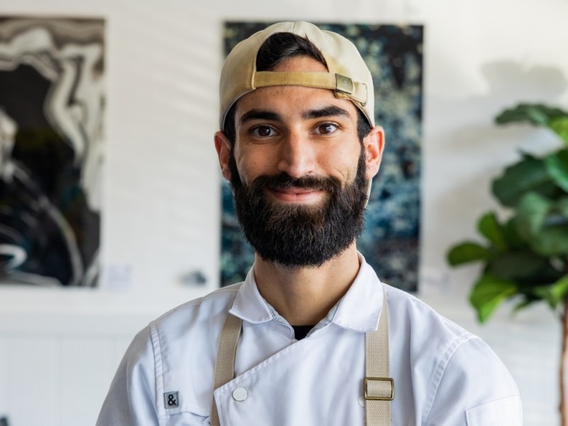 A close-up photograph of a smiling man with a beard and dark hair, wearing a white chef's coat with light tan suspenders and a light tan baseball cap worn backward. He is indoors, framed by abstract art on the wall and a large houseplant to his right.
