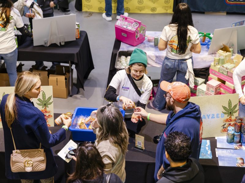 A vendor in a teal beanie pours a sample of a Dayzed beverage for an attendee at a crowded booth during the High & Dry Festival.