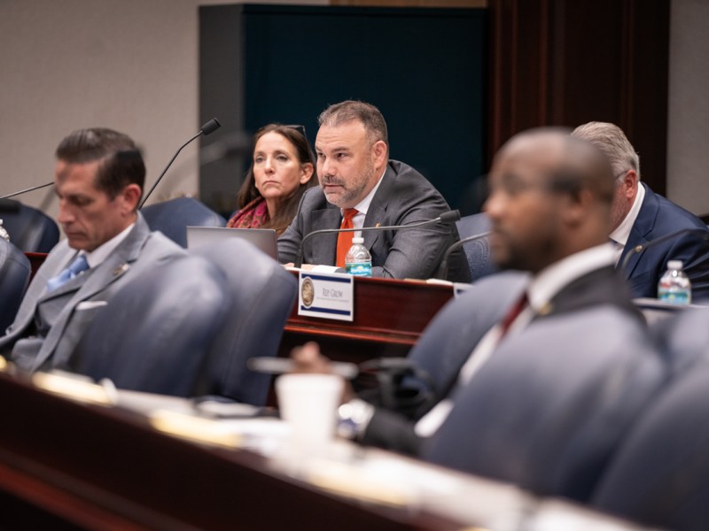 In a formal hearing room, several people are seated in rows. The central focus is on a man with a beard, wearing a grey suit and red tie, looking forward attentively.