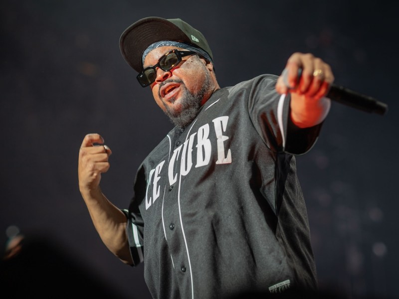 Ice Cube on stage, wearing a black jersey and sunglasses, holding the microphone out and gesturing with his left hand towards the crowd.