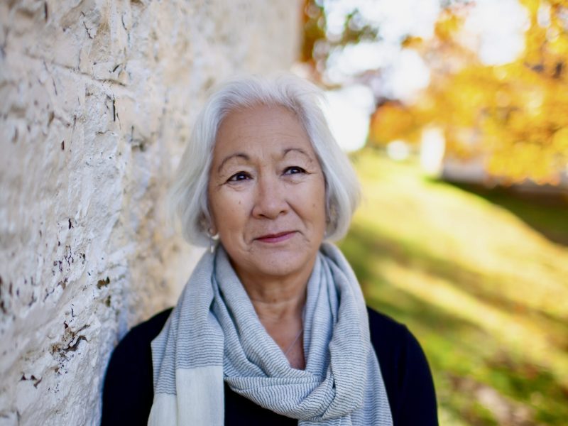 A portrait of an older woman with short white hair leaning against a textured stone wall. She wears a light grey scarf over a black top and looks off to the side with a calm, contemplative expression. The background is softly blurred, showing vibrant yellow and orange autumn foliage.