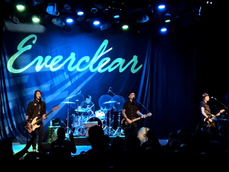 A rock band, Everclear, performs on a dark stage illuminated by blue and green lighting. A large, dark banner behind the band has the word "Everclear" written across it in a bright, light green script font. Four band members are visible: a bassist on the left, a drummer seated in the center, a guitarist/vocalist in the center-right, and another guitarist/vocalist further right. Silhouettes of the audience are visible in the foreground.