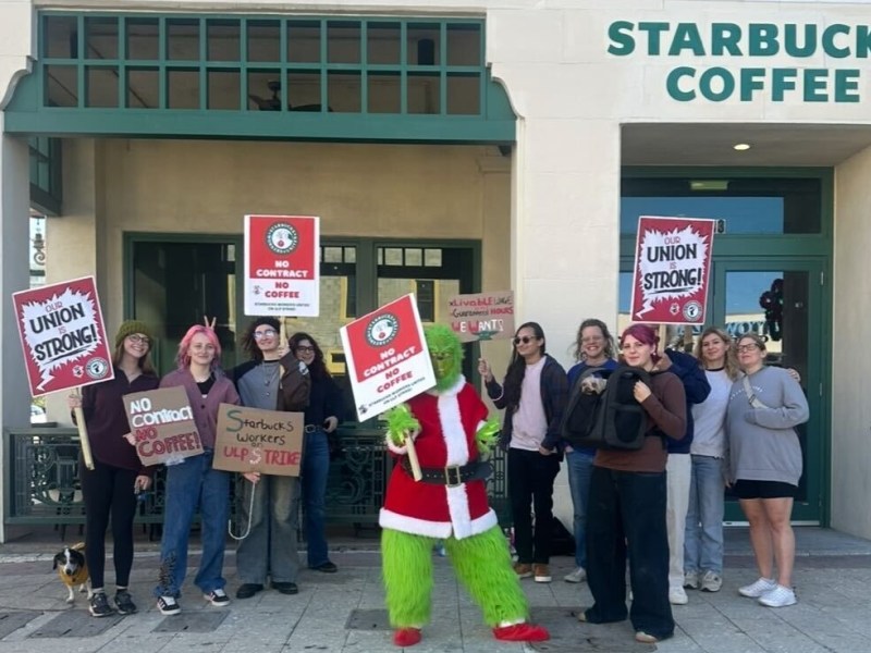 A group of people stands outside a "Starbucks Coffee" storefront holding red protest signs with the Starbucks Workers United logo. In the center, a person dressed as The Grinch in a Santa suit holds a sign reading "No Contract No Coffee," while others hold signs saying "Our Union Is Strong!" and "Starbucks Workers on ULP Strike". A handwritten sign in the background demands a "Livable Wage" and "Guaranteed Hours".