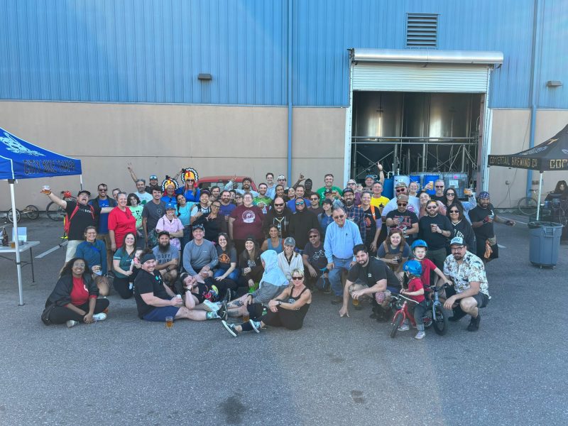 A large group photo of approximately 50 people gathered outdoors in front of an industrial building with blue siding. An open garage door behind the group reveals large stainless steel brewing tanks. To the left stands a blue tent labeled "City Bike Tampa," and to the right is a black tent for "Coppertail Brewing Co." The diverse crowd is dressed casually, with some holding beverages, and includes people sitting on the ground in the front row, next to a child on a small bicycle.
