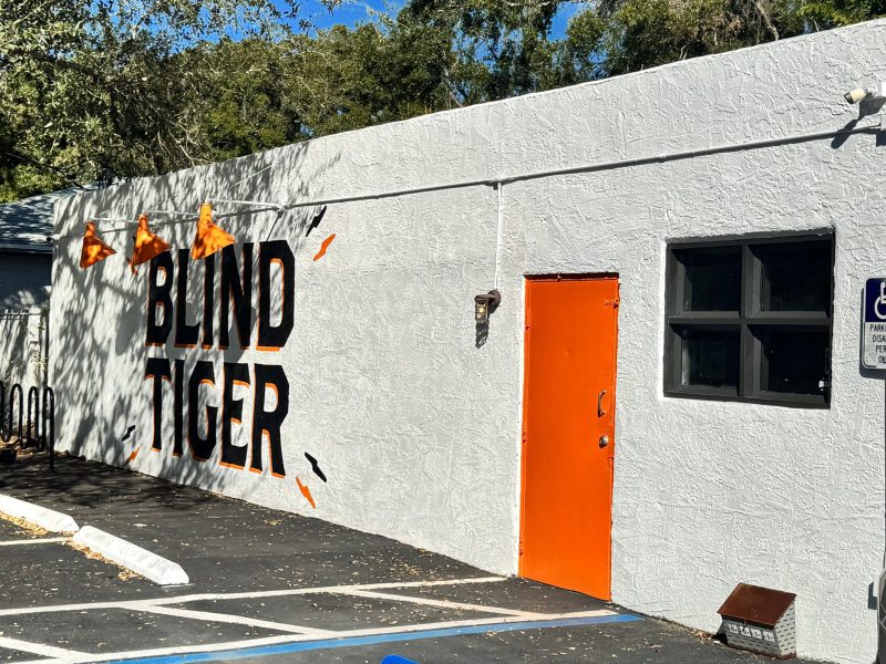 A street-level shot of the side of a white, textured building on a sunny day. The words "BLIND TIGER" are painted in large, black, stylized letters on the wall, with orange triangular banners strung on a wire above. To the right of the text is a bright orange door, a small black-framed window, and a blue "Parking by Disabled Permit Only" sign. A parking lot is in the foreground.