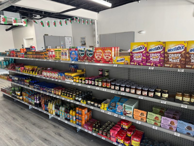 An interior view of a grocery store aisle. Grey shelves are stocked with various food products, including breakfast cereals like Weetabix and Alpen, jars of jam and condiments, bottled drinks, and baking supplies. A string of Welsh flag bunting hangs across the top of the aisle.