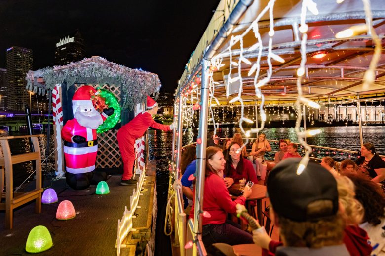A festive night scene aboard a tour boat decorated with hanging string lights. Passengers seated at tables look out toward a floating platform where a person in a red holiday costume stands next to a large inflatable Santa Claus and a small hut. The platform is lined with colorful, gumdrop-shaped lights, and a city skyline with illuminated skyscrapers is visible across the water in the background.