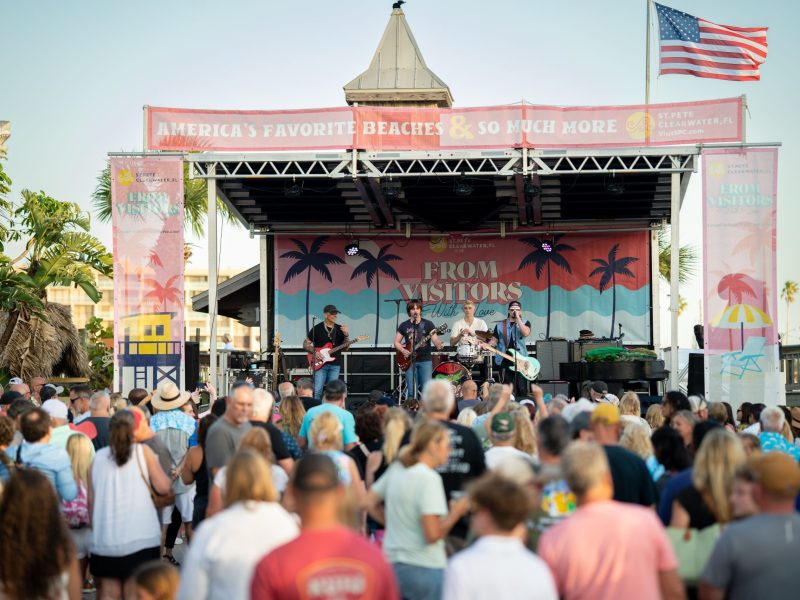 A low-angle, medium shot shows an outdoor concert with a band performing on a stage in front of a large crowd. Four musicians are on stage playing guitars and drums. Behind them, a large backdrop features palm trees and text reading "FROM VISITORS." A prominent red banner above the stage reads, "AMERICA'S FAVORITE BEACHES & SO MUCH MORE," with "ST PETE CLEARWATER" logos. An American flag flies on a pole to the right of the stage. The large audience in the foreground is seen from behind, facing the stage.