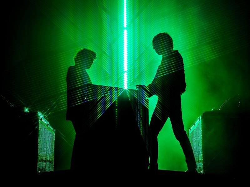 A striking, high-contrast silhouette of two people standing behind a large console or setup on a dark stage. A vibrant, vertical beam of bright green laser light runs down the center of the image, illuminating the air and creating a distinct, textured backdrop behind the figures. The green light also catches the edges of the console and equipment on either side, highlighting the concert setting.