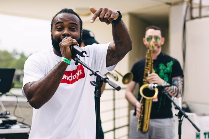 Rapper J.IN.X performs outdoors with a microphone in hand, pointing toward the crowd, while a saxophone player accompanies him in the background.