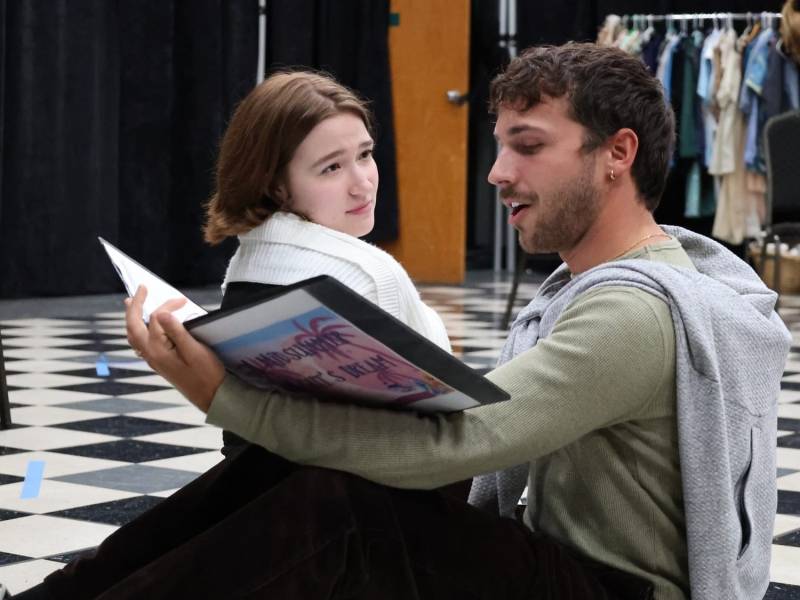 Two young actors sit on a black and white checkered floor during a rehearsal, with the man speaking while holding a script and the woman looking at him with a quizzical expression.