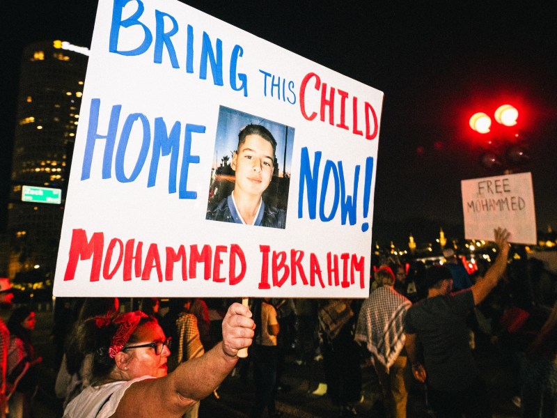 Nighttime protest scene where a woman holds a large sign that reads "BRING THIS CHILD HOME NOW! MOHAMMED IBRAHIM" with a photo of the boy in the center. Other protesters and city lights are visible in the background.