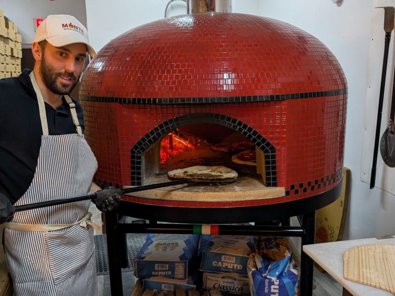 A pizza chef wearing a striped apron and a cap with the "Monte" logo stands next to a large, domed, red-tiled wood-fired oven. He holds a long peel with a pizza on it near the oven's open mouth, where glowing embers and another cooking pizza are visible inside. Bags of flour are stacked beneath the oven stand, and piles of pizza boxes line the wall in the background.